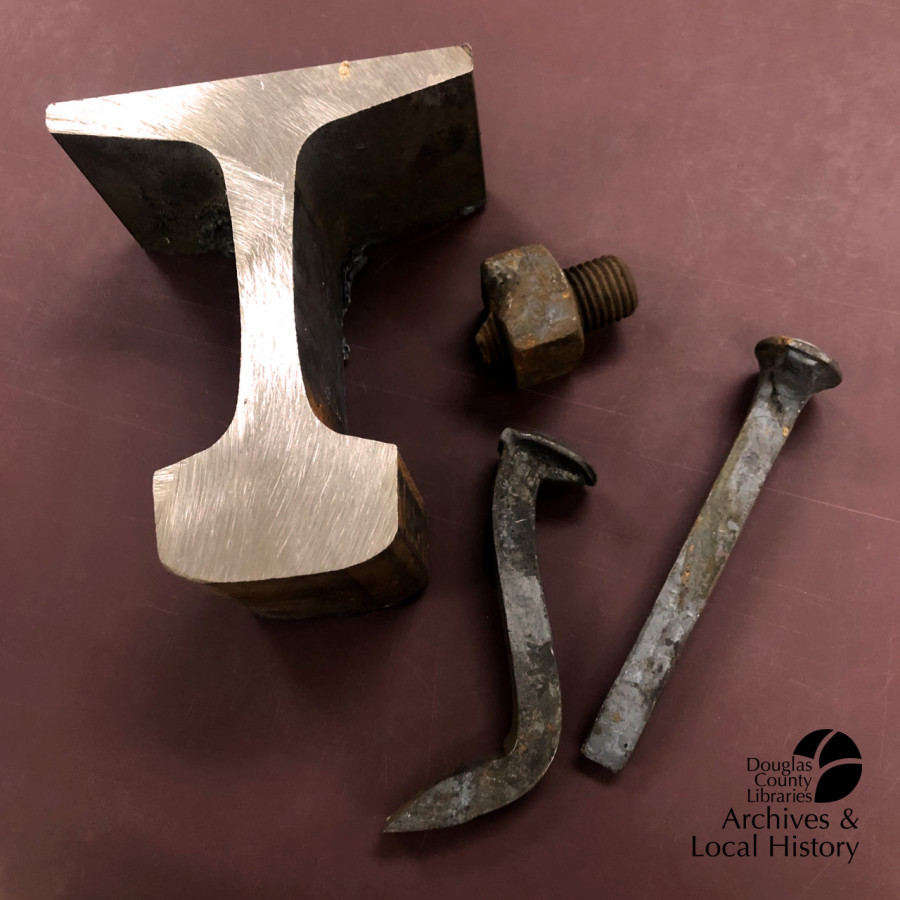 Image shows railroad irons including a piece of track, a bolt, and two spikes.