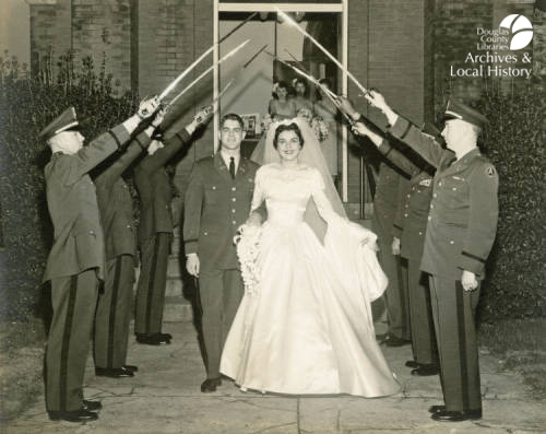 Image shows bride Joan Cooley and groom Andrew Cooley leaving their wedding ceremony under an arch formed by the sabers of an Honor Guard. This is the winner of the Archives Award for Best Wedding Dress.